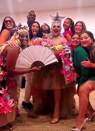Colorful group photo at an indoor celebration featuring a central drag performer in a star headpiece and sparkly makeup, surrounded by friends in floral costumes and bright accessories, one holding a large pink folding fan — festive party vibe.