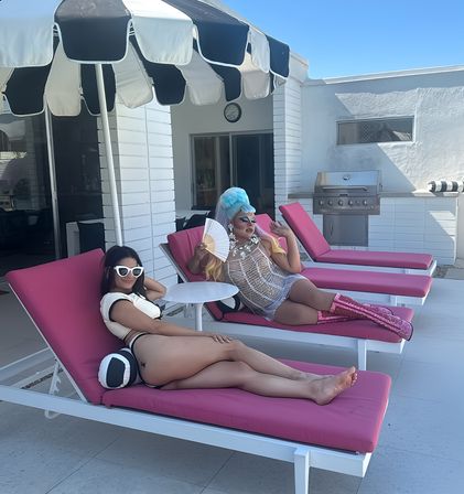 Sunny modern poolside patio with two people lounging on bright pink chairs under a black-and-white striped umbrella — one in a blue wig, sheer dress and pink sequined boots fanning themself, the other in white sunglasses and a bikini near a stainless steel grill.