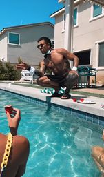 Playful backyard pool party: shirtless man in sunglasses, bow tie and cuffs crouches at the pool edge offering a red shot cup to someone in the water, turquoise pool, house and patio furniture in the sunny background.