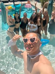 Summer backyard pool party selfie — person in pink heart sunglasses and bow tie poses in foreground while a cheering group of friends in swimsuits gathers in the turquoise pool.
