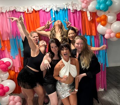 Seven women celebrating at an indoor party photo booth, striking playful poses in front of a pink, orange and blue fringe backdrop with white and colorful balloon garlands.
