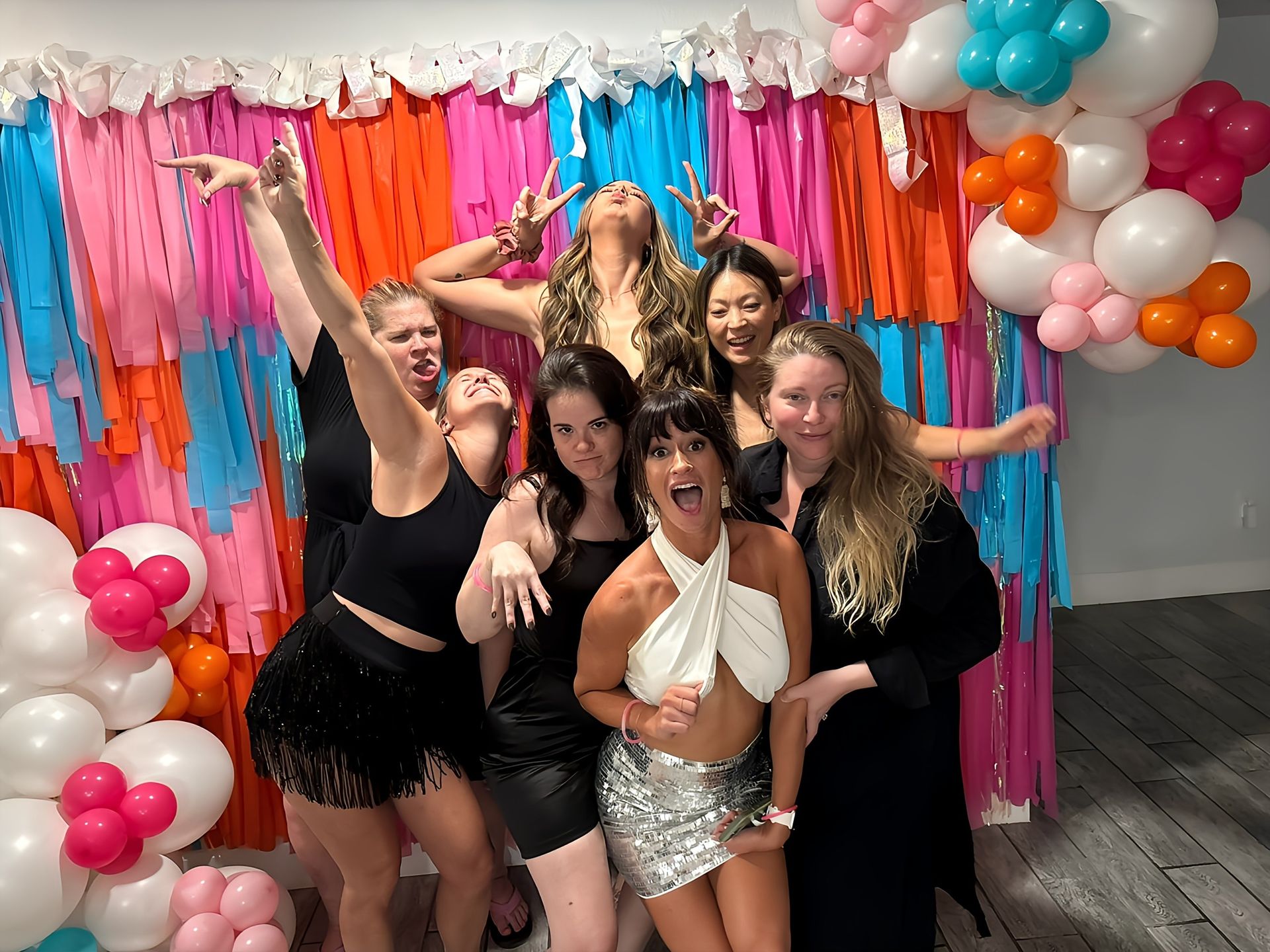 Seven women celebrating at an indoor party photo booth, striking playful poses in front of a pink, orange and blue fringe backdrop with white and colorful balloon garlands.