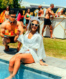 Bachelorette pool party at a sunny backyard pool — bride-to-be in a white robe and heart-shaped sunglasses dips her feet while shirtless servers bring colorful cocktails and guests mingle in swimwear.