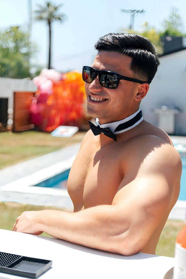 Smiling shirtless man in sunglasses and a black bow tie leaning poolside at a sunny backyard summer pool party.