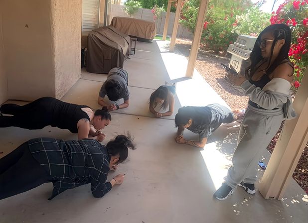 Five people doing a plank workout on a sunny backyard patio under a covered porch—four holding forearm planks while one stands with a phone near a BBQ grill and red flowering shrubs.