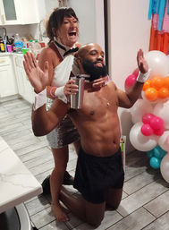 Playful kitchen party scene with a laughing woman in a white halter top and sequin skirt behind a kneeling, bearded shirtless man wearing cuffs and a bow tie, holding a tumbler next to a colorful balloon column.