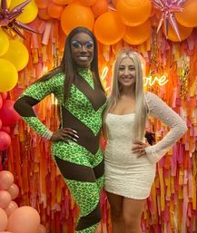 Two partygoers pose in front of a vibrant orange-pink fringe and balloon photo wall at a celebration — one in a green leopard-print and black sheer bodysuit with bold stage makeup, the other in a white sequin mini dress and tiara with a neon sign glowing behind them.