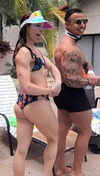 Poolside scene on a tropical pool deck: fit woman in floral bikini and colorful visor playfully sticking out her tongue, beside a man in black swim trunks with sunglasses and a sleeve tattoo, lounge chairs and palm tree in background.