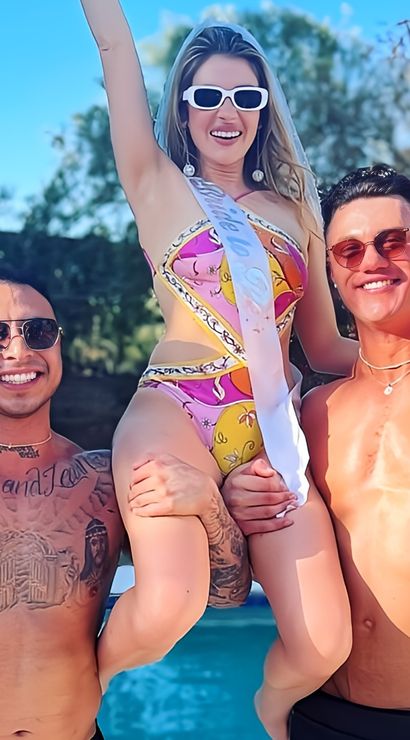 Sunny backyard pool party with a woman in a colorful swimsuit and bridal sash lifted and cheering between two shirtless, flexing men in sunglasses.