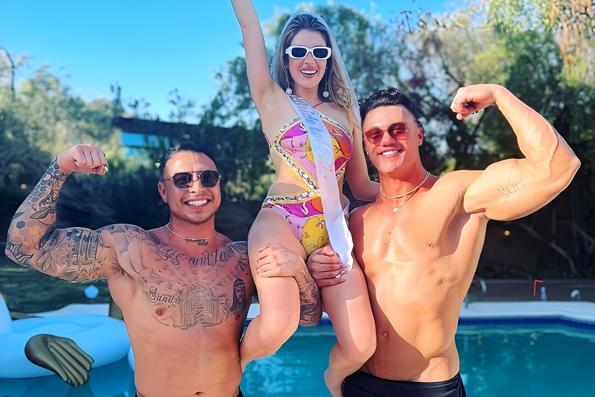 Sunny backyard pool party with a woman in a colorful swimsuit and bridal sash lifted and cheering between two shirtless, flexing men in sunglasses.