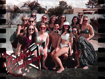 Group of friends in swimsuits posing at a sunny backyard pool party with palm trees and a white wall, smiling for a playful film-style group photo.