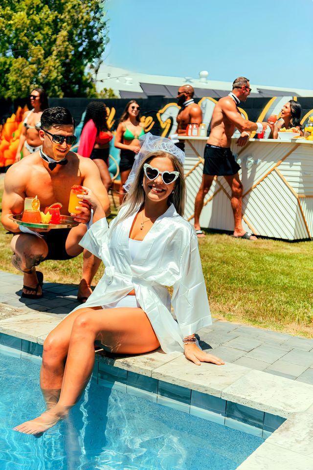 Bachelorette pool party by a sunny backyard pool — smiling bride-to-be in a white robe, veil and heart sunglasses sits with feet in the water as shirtless servers bring colorful cocktails and guests mingle by a poolside bar.