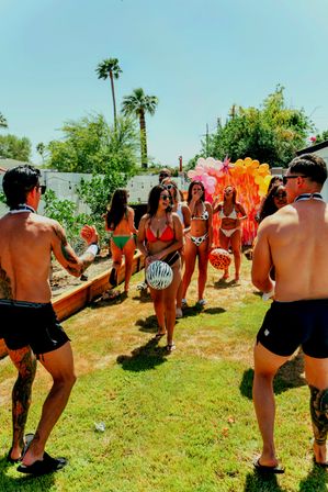 Friends in swimsuits playing with inflatable beach balls in a sunny backyard with palm trees and a colorful balloon arch