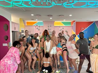 Large diverse group of friends in colorful swimwear laughing and posing in a vibrant vacation-rental kitchen with neon geometric ceiling lights, pastel cabinets, and bold mural — lively indoor party vibe