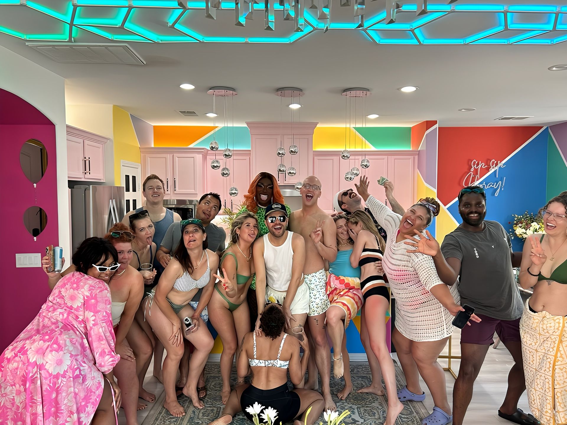 Diverse group of adults in swimwear laughing and posing in a bright, colorful modern kitchen with pastel pink cabinets, geometric neon ceiling lighting and hanging disco-ball ornaments — playful indoor party scene.