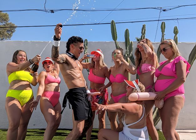 Group of women in bright pink and neon swimsuits laughing as a shirtless entertainer sprays champagne at a sunny backyard party with tall cacti against a white wall.
