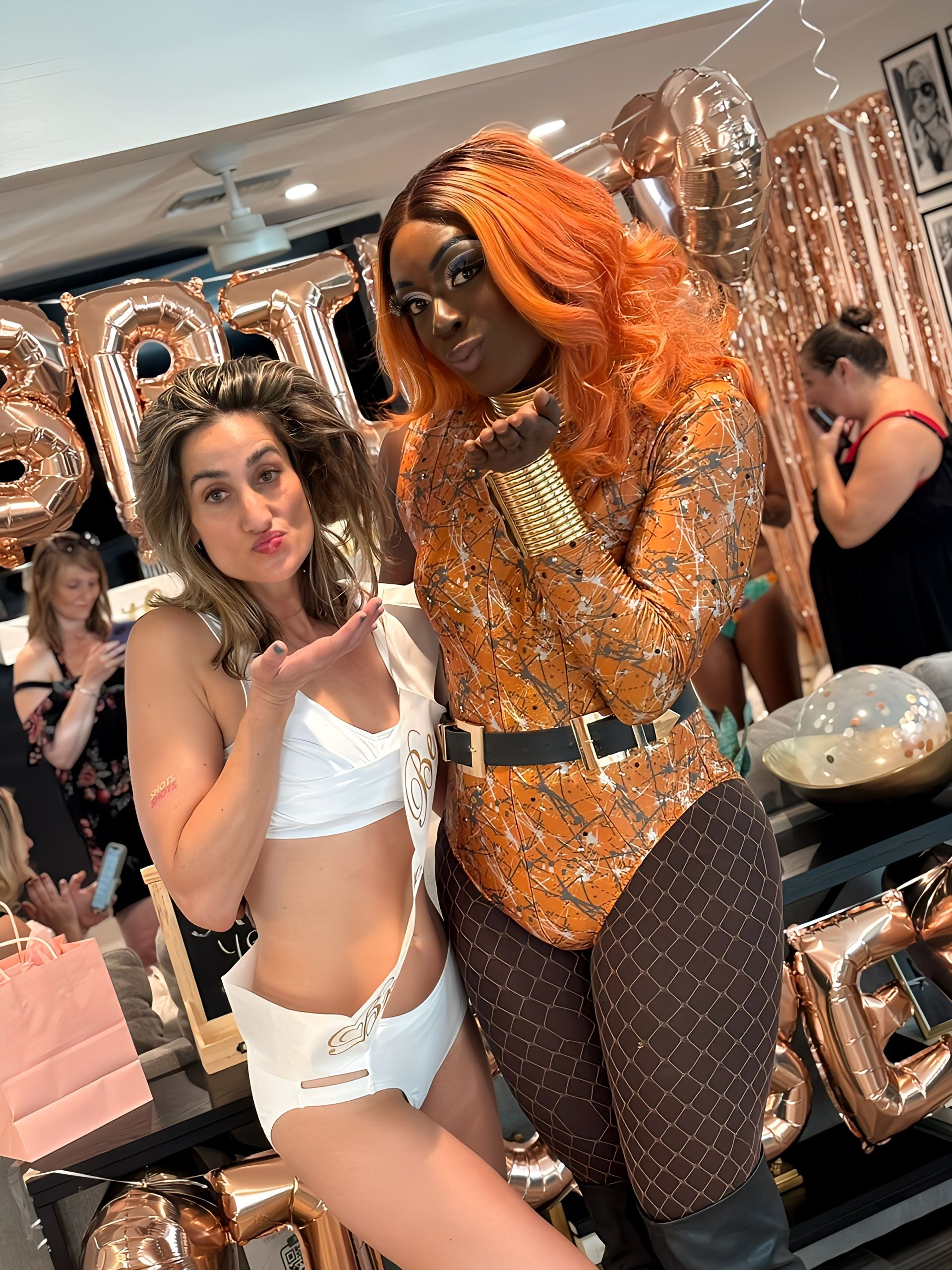 Two people at an indoor bachelorette party posing in front of rose-gold balloons — guest in white sash and swimsuit and performer in an orange wig and patterned bodysuit blowing a kiss.