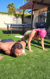 Two people doing push-ups on artificial turf in a sunny palm-lined backyard patio with a hanging egg chair and ping-pong table.