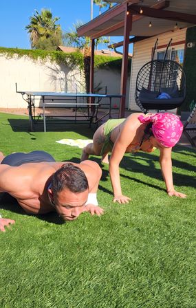 Two people doing push-ups on artificial turf in a sunny palm-lined backyard patio with a hanging egg chair and ping-pong table.