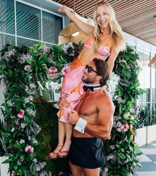 Smiling woman in a pink bikini and sarong lifted by a shirtless man wearing a bow tie and cuffs in front of a lush floral arch on a sunny rooftop patio — playful summer party photo-op.