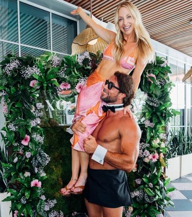 Smiling woman in a pink bikini and sarong lifted by a shirtless man wearing a bow tie and cuffs in front of a lush floral arch on a sunny rooftop patio — playful summer party photo-op.