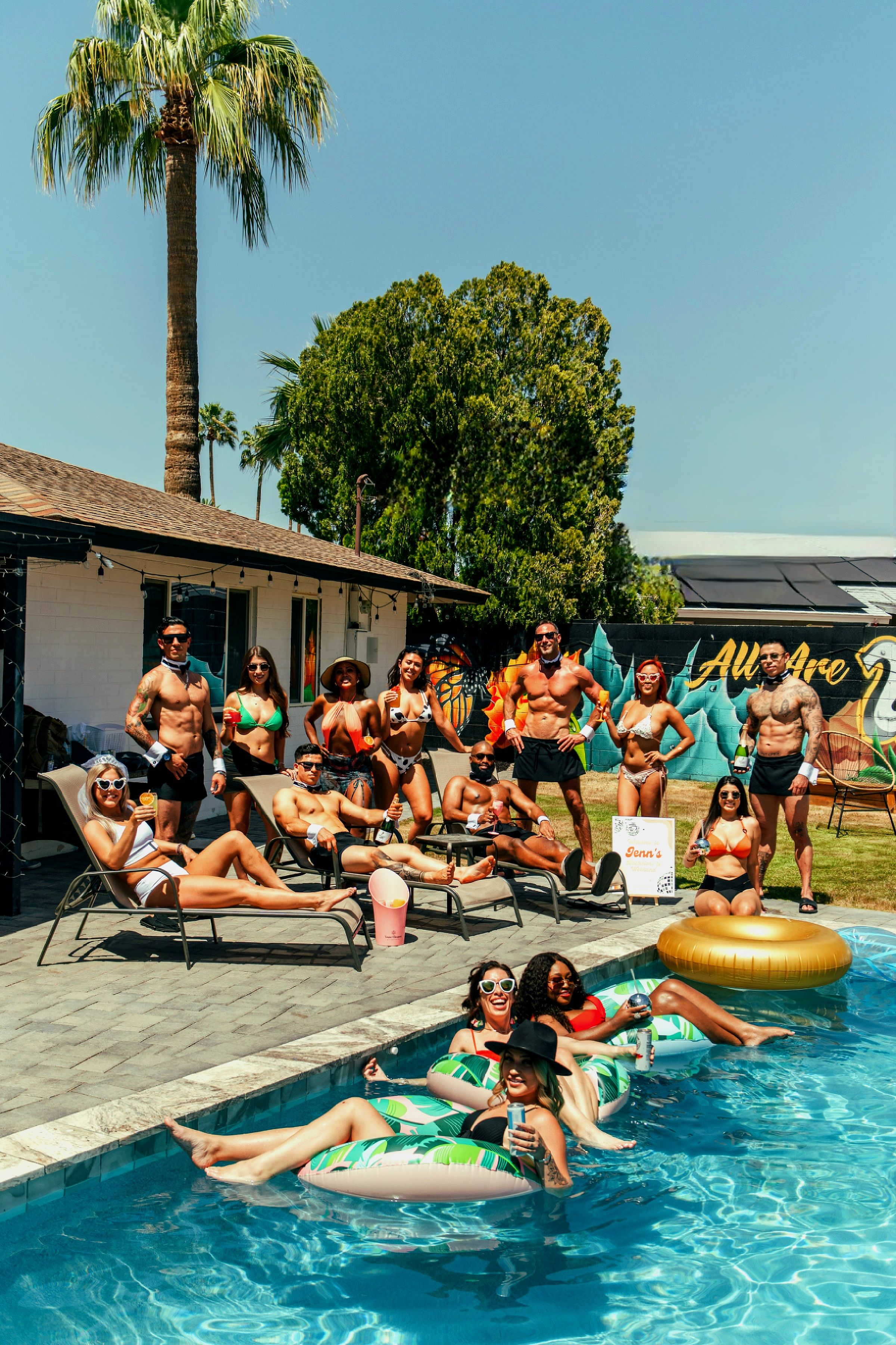 Lively backyard pool party on a sunny day with palm trees and a colorful mural, friends in swimsuits lounging on chairs and inflatable floats, laughing and sipping drinks by the pool.