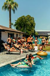 Sunny backyard pool party under tall palm trees — lively group in swimwear lounging on sun chairs and floating on inflatable tubes with drinks, colorful mural on the fence in the background.