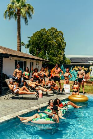 Sunny backyard pool party under tall palm trees — lively group in swimwear lounging on sun chairs and floating on inflatable tubes with drinks, colorful mural on the fence in the background.