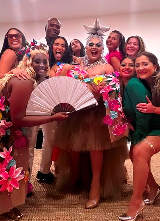 Lively group photo at an indoor tropical-themed party featuring a joyful drag performer in a star crown and jeweled makeup holding a large folding fan, surrounded by friends in colorful floral costumes and bright smiles.