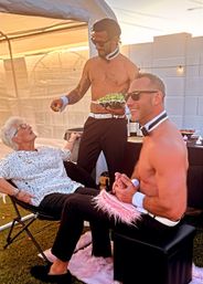 Outdoor sunset garden party with two shirtless male entertainers in bow ties and cuffs offering a bowl of grapes to a laughing seated woman under a white tent on a lawn.