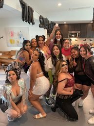 Smiling group of women in playful costumes posing at a decorated living-room costume party with black balloon banner and kitchen in background
