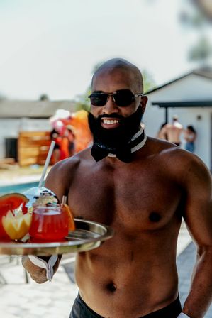 Smiling shirtless man in sunglasses and a bow tie collar serving colorful tropical cocktails on a tray at a sunny backyard pool party