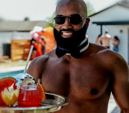 Shirtless server wearing sunglasses, bow tie and cuffs smiling poolside while carrying a tray of colorful cocktails at a backyard pool party