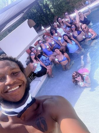 Smiling man in a bow tie takes a selfie at a sunny backyard pool party with a large group of women in colorful swimwear lounging in the shallow pool and cabana, each holding sparkling cocktails.