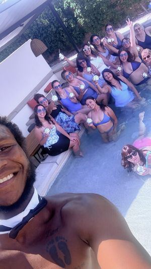 Smiling man in a bow tie takes a selfie at a sunny backyard pool party with a large group of people in swimsuits holding frozen cocktails under a pergola.