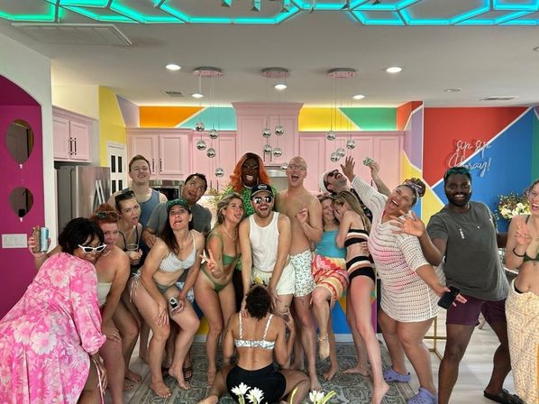 Large group of people in swimwear posing playfully in a bright pastel kitchen with geometric wall colors, hanging metallic pendant lights and neon ceiling accents — lively indoor pool-party scene.
