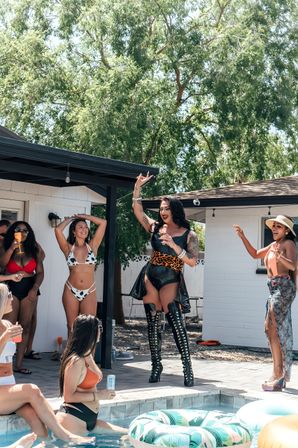 Backyard pool party with a costumed performer in black latex, leopard-print belt and thigh-high lace-up boots entertaining friends in swimsuits beside a pool with colorful inflatable rings and leafy trees overhead.