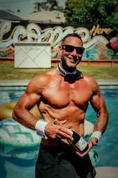 Shirtless man in sunglasses with bow tie cuffs opening a champagne bottle by a sunny backyard pool, colorful mural and inflatable floats in the background — playful pool party vibe.