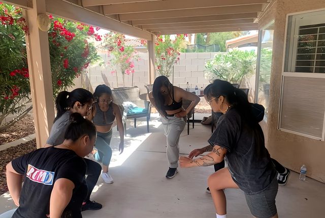 Group dance practice on a shaded suburban backyard patio — five people in activewear stretching and rehearsing under a pergola with flowering shrubs, wicker chairs and a water bottle nearby.
