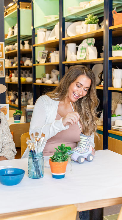 Two smiling people painting pottery at a bright paint-your-own ceramics studio, surrounded by shelves of colorful plates, brushes, and craft supplies.