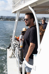Man in sunglasses holding a canned drink, leaning on the railing of a boat during a sunny coastal bay cruise with waterfront city skyline and other passengers in the background.