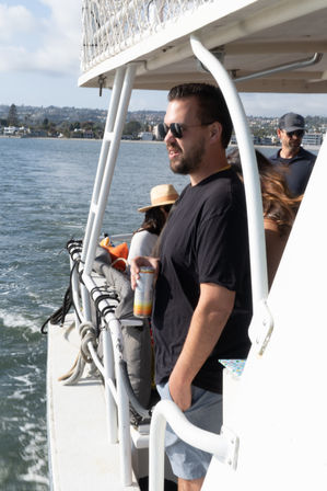 Man in sunglasses holding a canned drink, leaning on the railing of a boat during a sunny coastal bay cruise with waterfront city skyline and other passengers in the background.