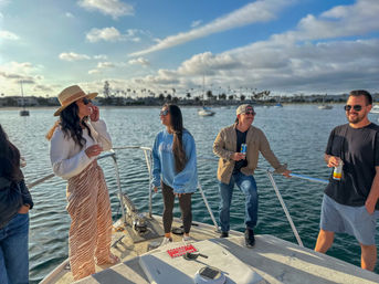 Group of four people laughing and holding drinks on the bow of a boat in a sunny coastal harbor with sailboats, palm trees and a blue sky with scattered clouds.