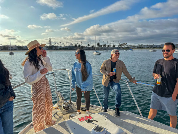 Group of four people laughing and holding drinks on the bow of a boat in a sunny coastal harbor with sailboats, palm trees and a blue sky with scattered clouds.