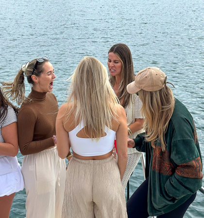 Four young women in casual summer outfits laughing and making faces together on a waterfront dock with calm blue water in the background