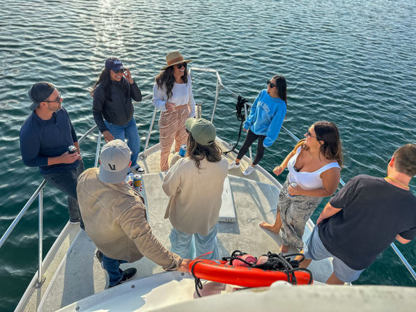 Group of friends on the bow of a boat over calm blue water, wearing casual summer clothes and sunglasses, chatting and holding drinks on a relaxed coastal outing.