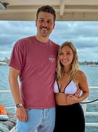 Smiling couple on a boat deck by the water — man in a maroon t‑shirt and jeans with arm around a woman in a white bikini top and black skirt, cloudy coastal skyline in the background.