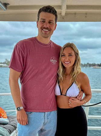 Smiling couple on a boat deck by the water — man in a maroon t‑shirt and jeans with arm around a woman in a white bikini top and black skirt, cloudy coastal skyline in the background.