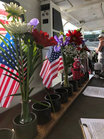 Patriotic display of red, white and purple flowers with small American flags in glass vases lined along a boat deck counter, papers and a coffee cup nearby at a marina