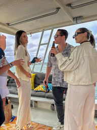 Friends chatting and sipping drinks on a covered boat deck in coastal waters — casual summer outfits, colorful cushions and ladder visible against a cloudy sky backdrop